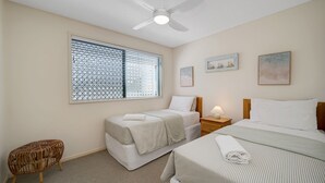 A bedroom with two beds, a ceiling fan, a window with blinds, and two wall-mounted paintings. - Seacrest Unit 2 Moffat Beach QLD (MOFFAT BEACH)