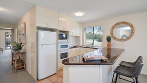 A kitchen with white cabinets, a refrigerator, a microwave, and a stove. There is a window with blinds, a round mirror, and a small table with a plant. - Seacrest Unit 2 Moffat Beach QLD (MOFFAT BEACH)