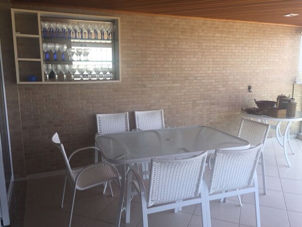 A dining area with a glass-top table and wicker chairs, a brick wall, and a shelf with wine glasses.