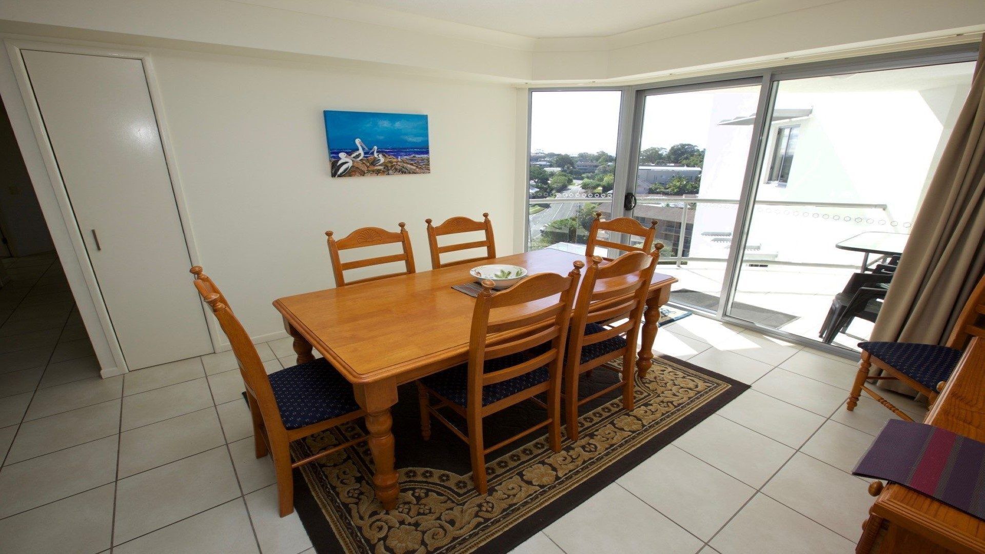 A dining area with a wooden table and chairs, a painting on the wall, and a view through a glass door.