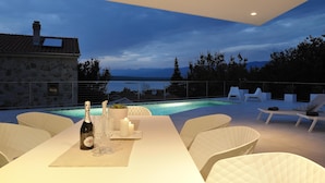 A dining area with a table set for two, a bottle of champagne, and candles. There is a pool in the background with lounge chairs around it. - Luxury Modern Seaview Villa  With  Private Pool In Klimno ***** (Klimno)