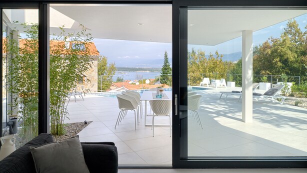 A modern living room with a large sliding glass door leading to an outdoor patio area with a pool, seating, and a view of a lake and mountains. - Luxury Modern Seaview Villa  With  Private Pool In Klimno ***** (Klimno)