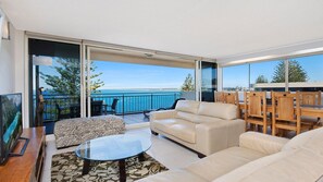 A modern living room with a sofa, coffee table, and a TV on a wooden stand. There is a balcony with a view of the sea and trees. - Wickham Point Unit 5 Kings Beach QLD (KINGS BEACH)