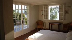 A bedroom with a bed, a chair, a small table, and a window with a view of greenery.