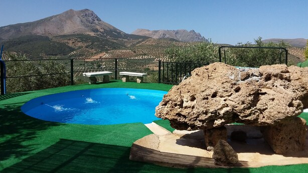 A pool with a rocky feature in the foreground, surrounded by a green lawn and a metal fence. In the background, there are mountains and a clear sky. - LA ALMORZARA BALCONY (Priego de Córdoba)