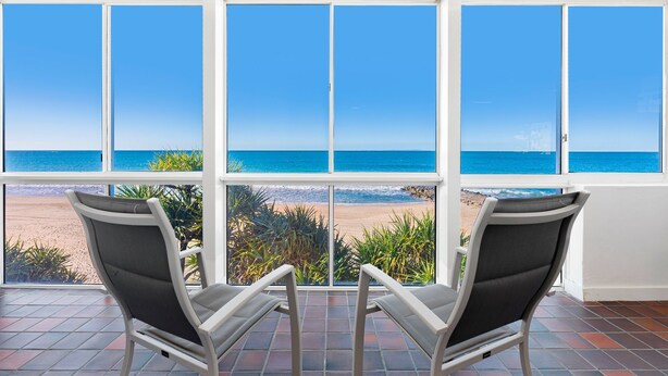 Una habitación con un ventanal grande con vista a una playa, dos sillas orientadas hacia la ventana y suelo de baldosas.