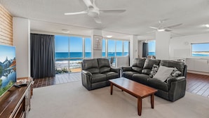 Un salón amplio con un gran ventanal con vistas al mar, sofás de cuero, una mesa de centro de madera y ventiladores de techo.