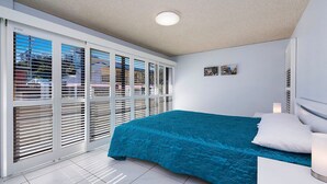 A bedroom with a bed, white walls, a ceiling light, and a view through large windows with blinds.