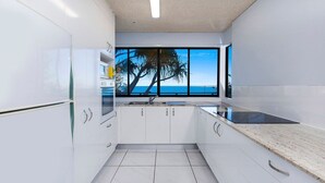 A modern kitchen with white cabinets, a black countertop, and a large window offering a view of palm trees and the sea.