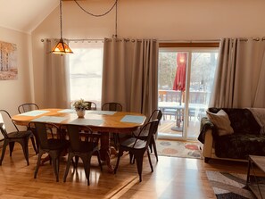 Un comedor con una mesa y sillas de madera, un jarrón con flores y vistas al exterior a través de amplias ventanas.