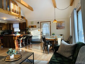 Una sala de estar acogedora con un sofá, una mesa de centro y un comedor con sillas y una mesa. Se observa una cocina con armarios de madera y una ventana.