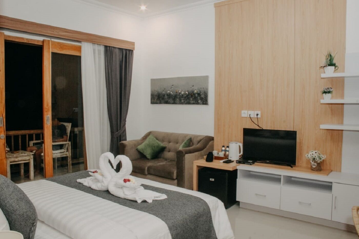 A modern hotel room with a bed, a sofa, a TV, and a sliding glass door leading to an outdoor area.