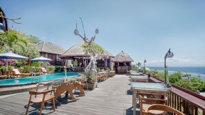 Une terrasse en bois avec une piscine, des chaises longues et des tables, offrant une vue sur l’océan.