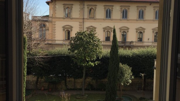 A view through a window of a building with a red-tiled roof, multiple windows, and a well-maintained garden with a tree and a stone pathway. - Fabulous 200sqm bright, new apartment walking distance to Duomo and city centre (Firenze)