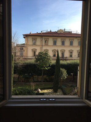 A view through a window of a building with a red-tiled roof, multiple windows, and a well-maintained garden with a tree and a stone pathway. - Fabulous 200sqm bright, new apartment walking distance to Duomo and city centre (Firenze)