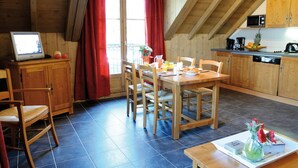 A cozy kitchen with wooden cabinets, a dining table with chairs, a small TV, and a window with red curtains. - Appartement Cosy 3 pièces | Ski, Sauna (Saint Gervais)