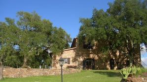 A stone house with a stone wall in front, surrounded by trees and a grassy lawn.