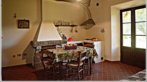A rustic kitchen with a tiled backsplash, wooden chairs, a table with a colorful tablecloth, and a hanging light fixture.