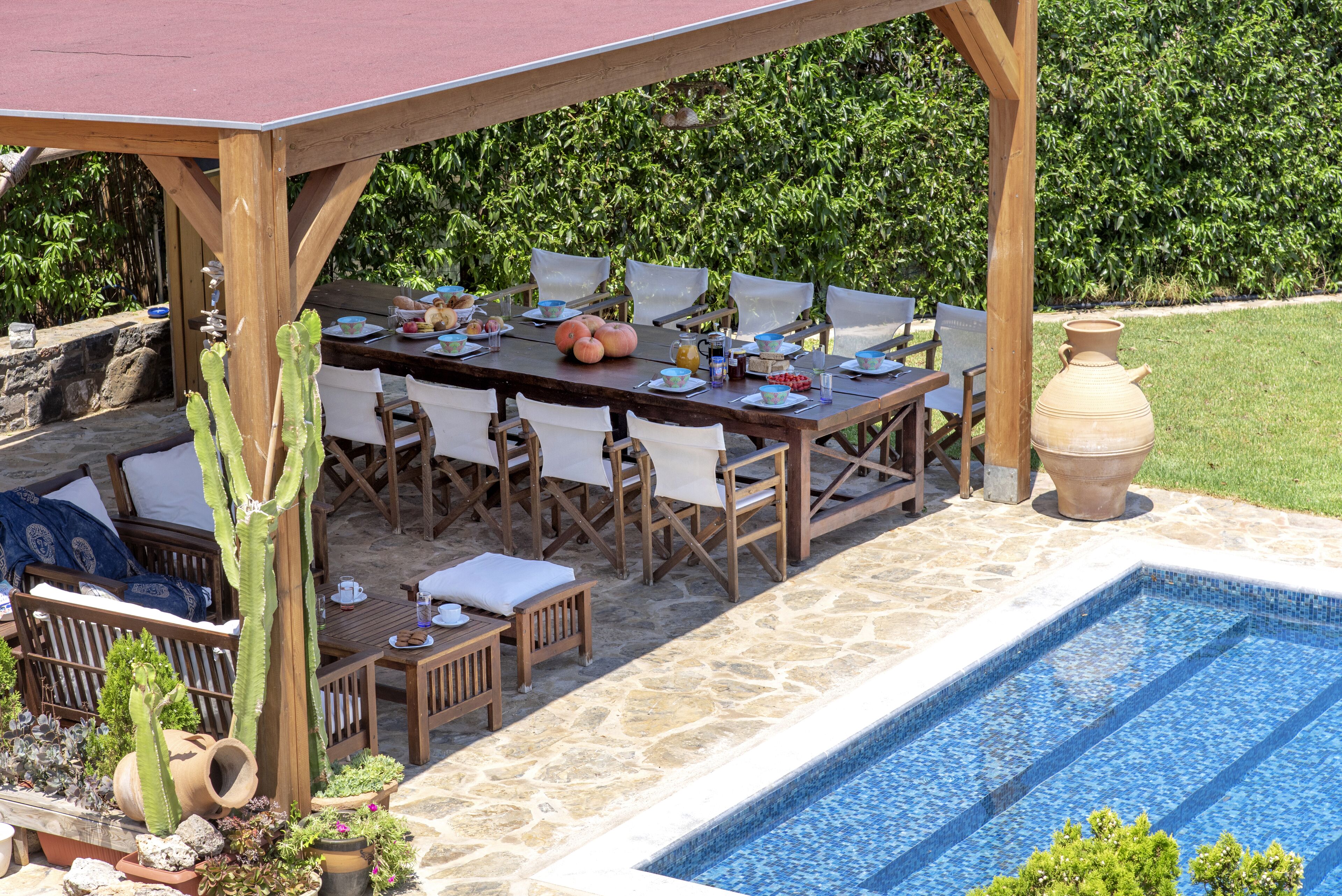 An outdoor dining area with a pool, wooden tables, chairs, and a pergola.