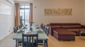 A dining area with a long table set for six, surrounded by chairs, a brown sofa, and a large abstract painting on the wall. - Bugibba - 2 bedroom apt 150m from Sea Front Fully Air Conditioned, Terraces WiFi (Bugibba,)