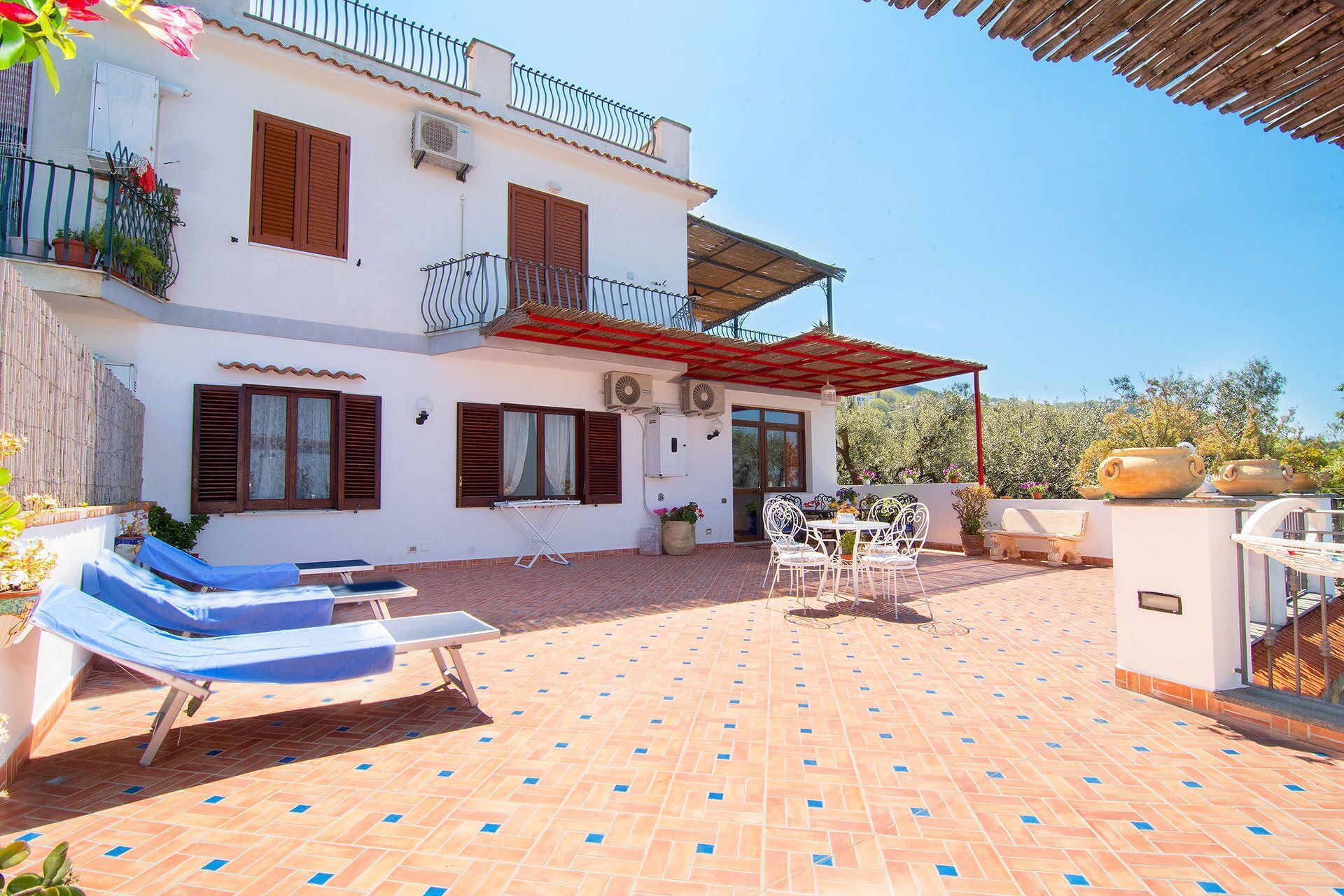 A white building with a tiled patio, outdoor furniture, and a red umbrella.