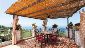 Ein überdachter Außenbereich mit Tisch und Stühlen, Blumentöpfen und Blick aufs Meer.