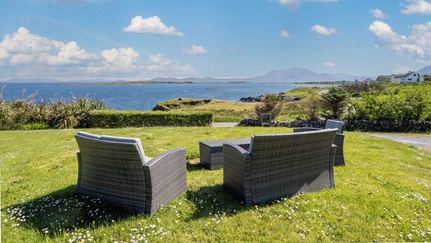A grassy outdoor seating area with wicker furniture overlooking a body of water and distant hills.