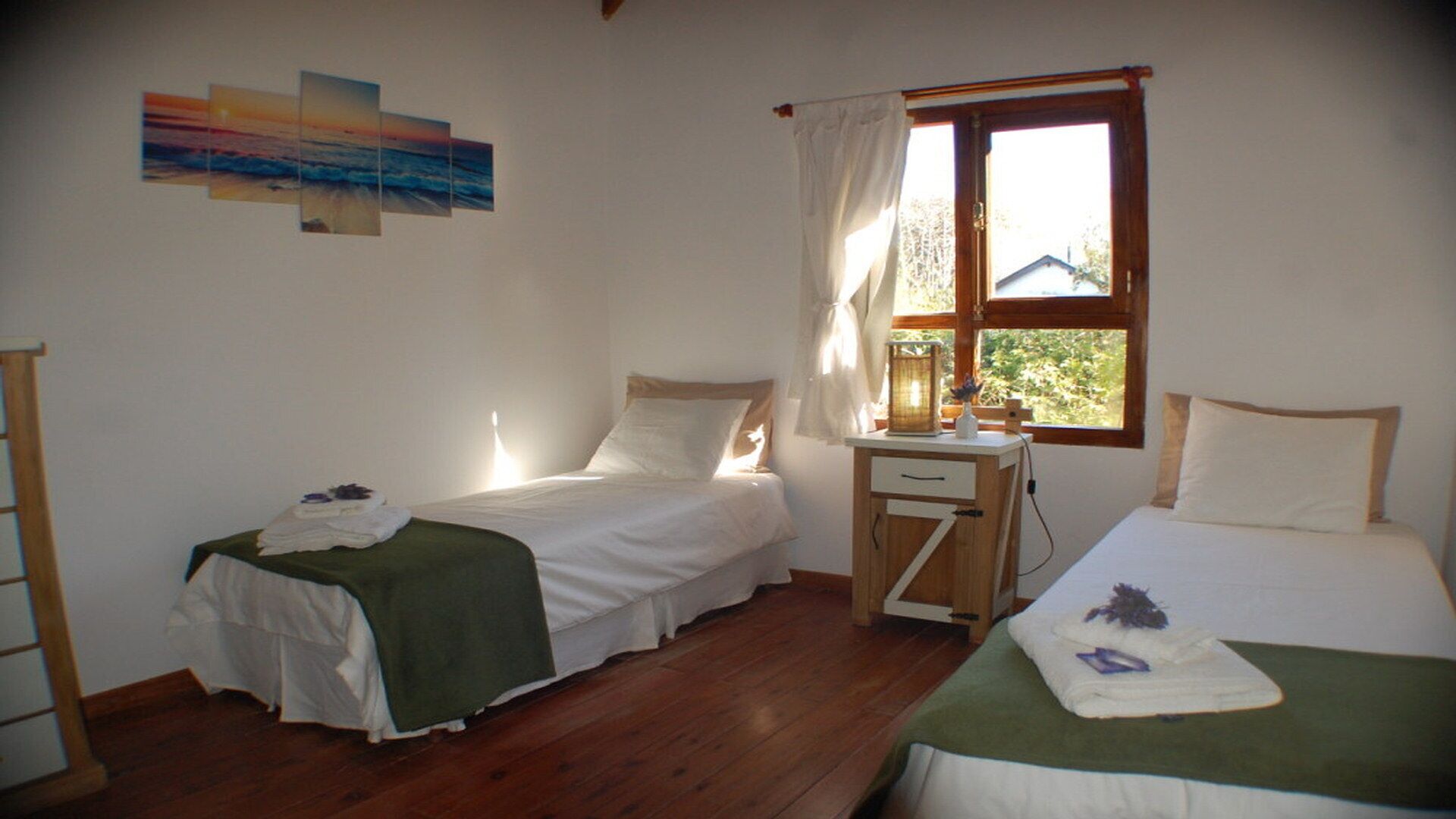 A room with two single beds, a small wooden nightstand, a window with curtains, and a painting on the wall.