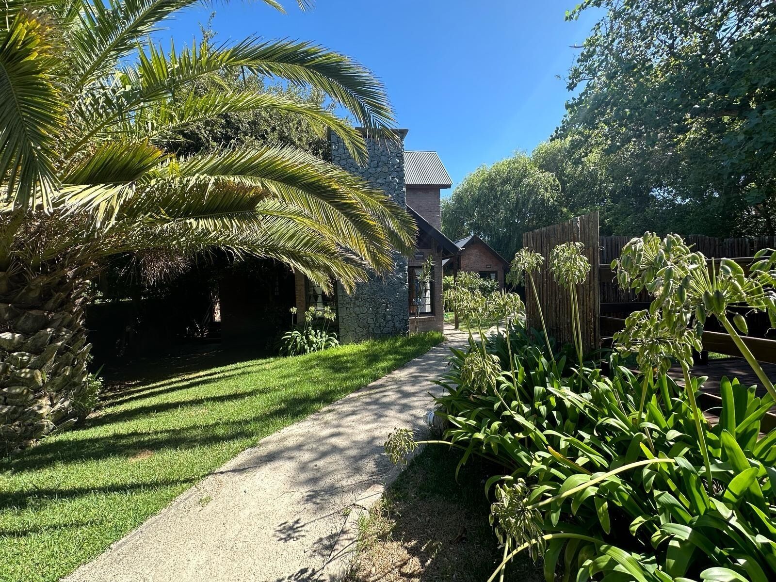 A pathway leading to a stone building with a brick roof, surrounded by lush greenery and a palm tree.