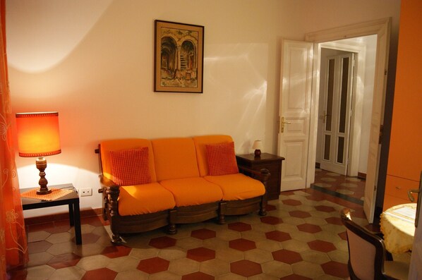 Une pièce comprenant un canapé, une table avec une lampe, un tableau encadré au mur et une porte donnant sur une autre pièce.