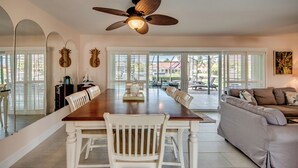 A spacious dining area with a wooden table and white chairs, a ceiling fan, and large windows with blinds. - West Facing Elec Heated Pool, 2024 Tritoon rental, Walk/Bike to Bars&Restaurants (Cape Coral)