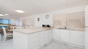 A modern kitchen with white cabinetry, a dining area with a table and chairs, and a view of the beach through large windows. - Kookaburra Court Unit 1 Bulcock Beach QLD (CALOUNDRA)