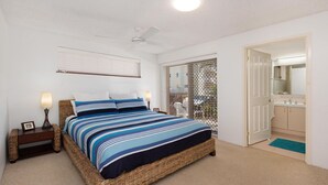 A bedroom with a bed, bedside lamps, a wicker nightstand, a ceiling fan, a window with blinds, and a bathroom with a sink and mirror. - Kookaburra Court Unit 1 Bulcock Beach QLD (CALOUNDRA)