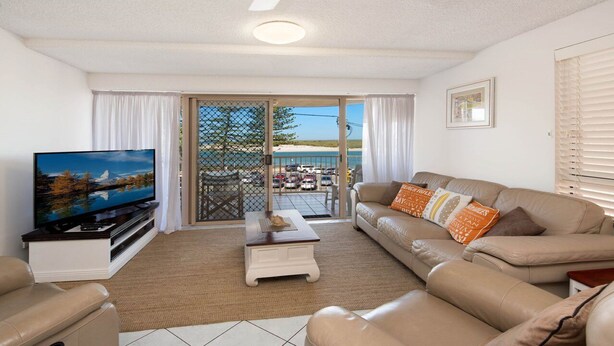 A living room with a television, sofa, and a view of a beach through a sliding glass door. - Kookaburra Court Unit 1 Bulcock Beach QLD (CALOUNDRA)