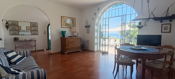 A spacious room with a wooden floor, a dining table with chairs, a sofa, a wooden cabinet, and a large arched window offering a view of the sea.