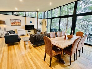 A modern living room with a sofa, TV, fireplace, dining table with chairs, and large windows offering a view of trees.