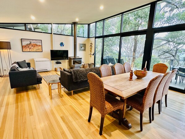Uma sala de estar moderna com sofá, televisão, lareira, mesa de jantar com cadeiras e janelas amplas com vista para árvores.