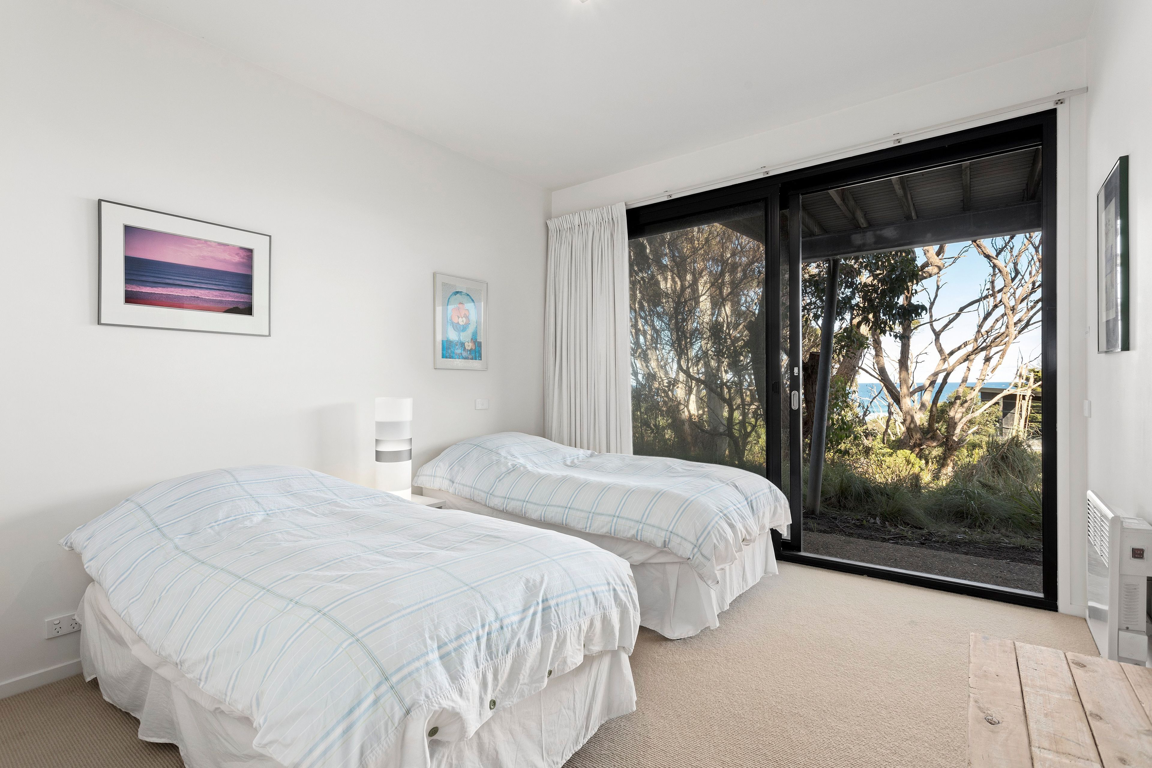 A bedroom with two beds, a large window with a view of trees, and artwork on the wall.