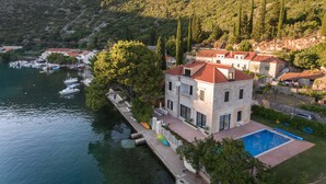 Una propiedad costera con piscina, un muelle y varias embarcaciones, y un cerro de fondo.