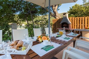 An outdoor dining area with a wooden table set for breakfast, including croissants, fruits, and beverages.