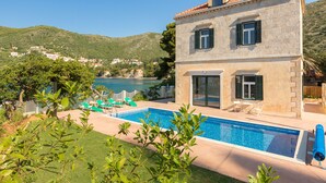 Un edificio de dos pisos con piscina, muebles de exterior verdes y vistas a un lago y colinas.