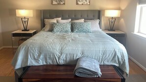 A neatly made bed with a grey and white patterned duvet, two matching pillows, and a folded grey towel on a wooden bench. There are two bedside tables with lamps, a headboard with a tufted design, and two framed pictures on the wall. - Beautiful Rehoboth Beach Condo! 3 bed 2 bath! 2 Pools! Tennis! Gym!  (Rehoboth Beach)