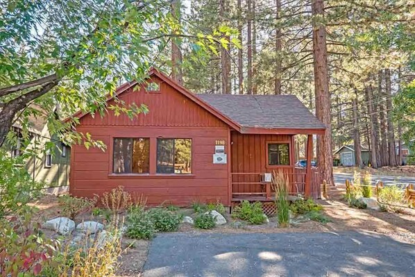 Une petite cabane en bois rouge, dotée d’un porche, entourée d’arbres et bordée d’une allée de gravier.