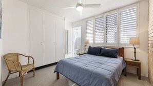 A bedroom with a bed, a chair, a nightstand, a lamp, and a window with blinds. - Jamarkos Unit 4 Kings Beach QLD (KINGS BEACH)