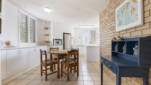 A kitchen with white cabinets, a wooden dining table, and chairs. There is a brick wall with a fish painting and shelves with jars. - Jamarkos Unit 4 Kings Beach QLD (KINGS BEACH)