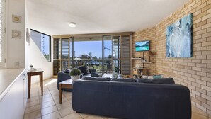 A modern living room with a large sofa, a TV mounted on the wall, and a view of the outdoors. - Jamarkos Unit 4 Kings Beach QLD (KINGS BEACH)