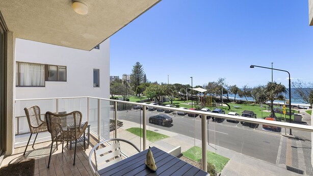 A balcony with wicker chairs and a table, overlooking a parking area and a view of the sea. - Jamarkos Unit 4 Kings Beach QLD (KINGS BEACH)