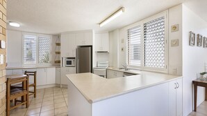 A modern kitchen with white cabinetry, a central island, a refrigerator, a microwave, and a stove. There are two windows with blinds, a hanging light fixture, and a small table with chairs. - Jamarkos Unit 4 Kings Beach QLD (KINGS BEACH)