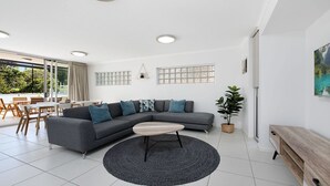 Ein modernes Wohnzimmer mit einer grauen Eckcouch, einem runden Couchtisch und einem Essbereich mit Holztischen und -stühlen.