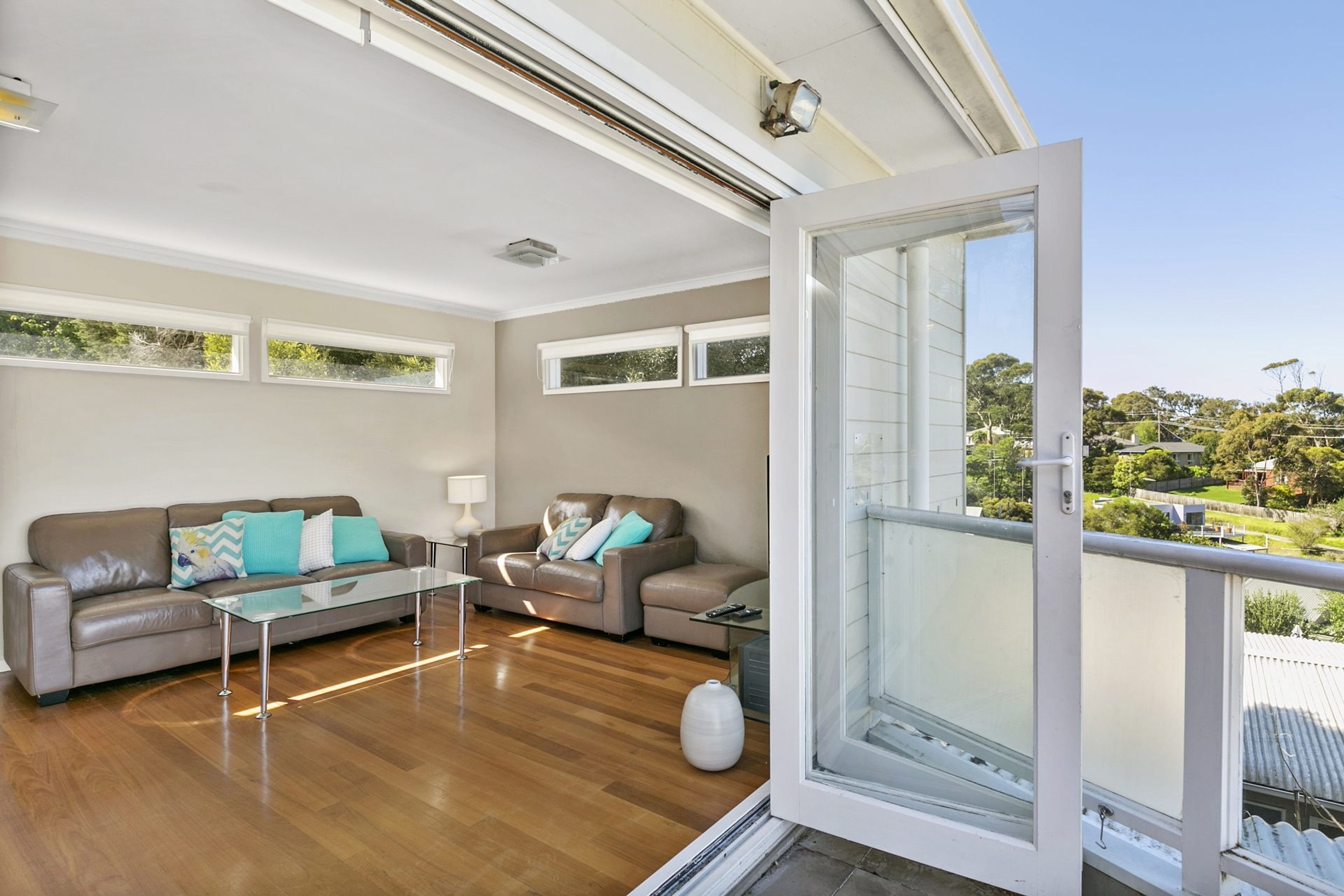 Un salon moderne avec une porte vitrée donnant sur un balcon, un plancher en bois et deux canapés en cuir avec des coussins.